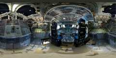 The Space Shuttle's flight deck - a panoramic view, showing the sheer number of controls and displays on walls and overhead panels. The blue squares lining the walls, overhead panels, and even backs of chairs are velcro pads used stop objects and things like checklists (cue cards) floating around when in space. The joystick in the top left corner is for the robotic arm, and the 2 nearby windows look on to the payload bay.